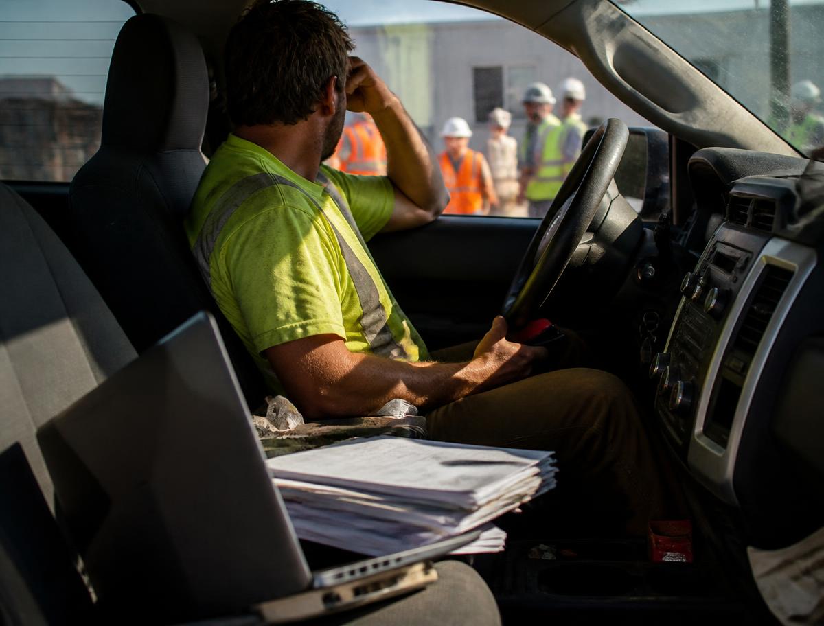 Frustrated contractor sitting in his pickup truck with paperwork and a laptop while his crew waits outside
